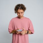 Adolescente de camisa rosa concentrado enquanto usa um smartphone diante de um fundo cinza.