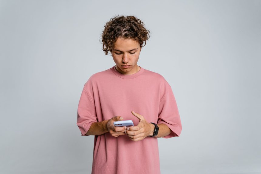 Adolescente de camisa rosa concentrado enquanto usa um smartphone diante de um fundo cinza.