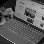 Laptop exibindo artigos de notícias, acompanhado de jornais e uma xícara de café sobre a mesa.