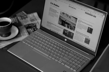 Laptop exibindo artigos de notícias, acompanhado de jornais e uma xícara de café sobre a mesa.