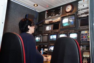 Profissional trabalha em uma sala de controle de TV com várias telas e equipamentos de transmissão.