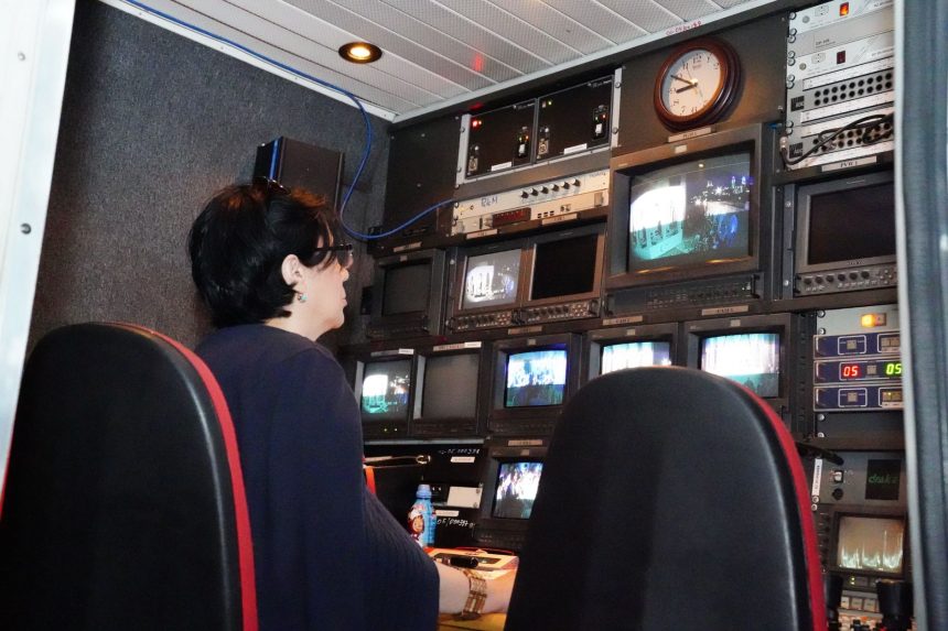 Profissional trabalha em uma sala de controle de TV com várias telas e equipamentos de transmissão.
