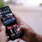 Mãos seguram um smartphone exibindo um vídeo ao ar livre.