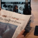 Mãos seguram um jornal ao lado de um laptop e um smartphone sobre uma mesa.