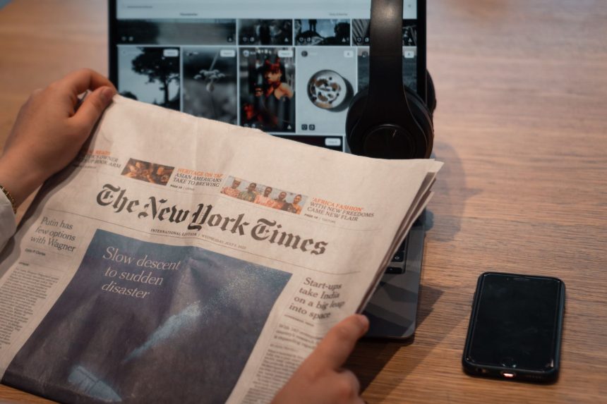 Mãos seguram um jornal ao lado de um laptop e um smartphone sobre uma mesa.