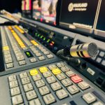 Painel de controle de um estúdio de transmissão com microfone e várias telas.