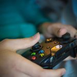 Mãos seguram um controle sem fio moderno de videogame em ambiente interno.