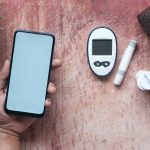 Mão segura um smartphone ao lado de um glicosímetro e materiais médicos sobre uma superfície de madeira