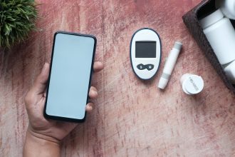 Mão segura um smartphone ao lado de um glicosímetro e materiais médicos sobre uma superfície de madeira
