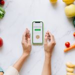 Mãos seguram um celular sobre uma bancada com frutas e verduras frescas.
