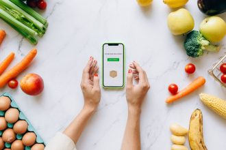 Mãos seguram um celular sobre uma bancada com frutas e verduras frescas.