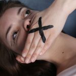 Mulher com a mão sobre a boca, simbolizando o silenciamento e a censura.